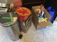 Photo showing large metal trash can, vintage red tin box on top of a cardboard box, cardboard box filled with aluminum foil roasting trays and wraps.