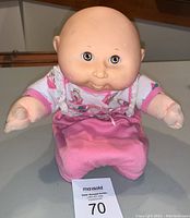 Full doll view showing soft pink outfit and the doll's bald vinyl head with wide eyes and plush arms and legs.