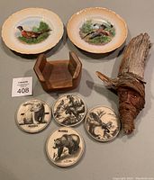 Full lot showing two vintage Germany pheasant plates, wooden coaster holder, four Alaska wildlife coasters, and hand-carved wooden man's face sculpture with live edge.