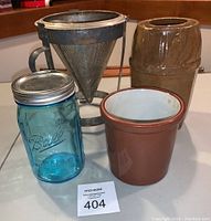 Full view of blue Ball canning jar, brown salt crock, and metal cone colander arranged together on table.
