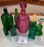 Full lot of a pink 1970 Ezra Brooks whiskey decanter with embossed design and matching pink glass stopper, surrounded by seven green vintage glass bottles in various sizes with different stoppers and plain tops.