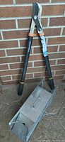 Fiskars lopper pruner with black and orange handles standing upright against a brick wall alongside the metal Havahart live animal trap on the ground.
