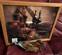 Oak framed print of a rustic still life with roasted pheasant, wine bottles, grapes, bread, and kitchen tools alongside a hand-painted wood duck decoy on a leather surface.