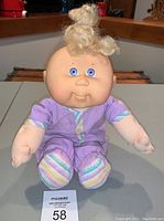 Front view of vintage 1989 Hasbro Cabbage Patch Baby Doll with curly blonde hair, blue eyes, and pastel purple outfit with striped trim.