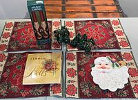 Photo of four rectangular tapestry poinsettia placemats arranged in two rows, 12 dark green Christmas tree napkin rings in front center, boxed pair of red Kurt Adler Santa taper candles upright on left. Items displayed on wooden surface.