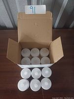 Open cardboard box containing 12 white plastic electric tea-light candles arranged in two rows of six, with six candles outside the box placed to show item details.