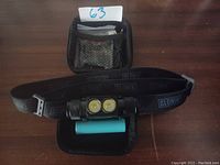 Top view of black battery powered headlamp with two LED lights, turquoise rechargeable battery, adjustable Slonik head strap, and small carrying case with mesh pocket.
