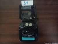 Black battery powered head lamp with dual LED lights, green rechargeable battery, USB charging cable, inside black zippered carrying case on wooden table.