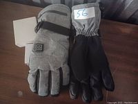 Photo showing pair of electric heated gloves, one grey fabric with power button, the other black palm
