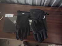 Photo showing both left and right black unpadded gloves with an adjustable wrist strap and what appears to be heating control features, displayed on a dark wooden surface with a black box underneath.