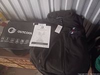 Outcool heated vest laid next to carrying bag and instruction manual on a wooden surface with metal background.