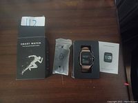 Photo showing smart watch box, USB charging cable, watch in plastic holder, and instruction manual.