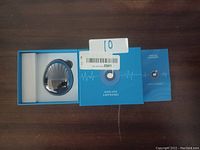 Wireless earphones in oval charging case alongside blue packaging box and user manual displayed on wooden surface.