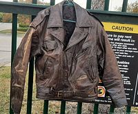 Brown leather Indian Motorcycle jacket hanging on a green fence, showing distressed leather and front details including zipper, pockets, and belt strap.