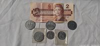 Photo shows one Canadian $2 bill from 1986 and six coins including four $1 coins from 1968, 1976 (two coins), 1979, and two 50 cent coins from 1974 and 1979.