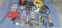 Overview of the lot showing multiple hand and power tools arranged on a blue tarpaulin outdoors including cordless drills, hammers, saw, extension cords, and various hand tools.