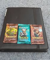 Three Magic The Gathering booster packs laid on a black binder, showing front packaging with artwork and branding for Strixhaven: School of Mages (two packs) and Theros Beyond Death (one pack).
