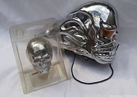 Photo showing large chrome skull headlight cover with orange eye lenses and smaller chrome skull accessory in plastic packaging.