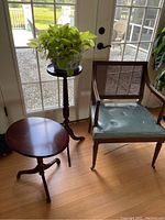 Set of three items: armchair with leather seat on casters, tall wooden plant stand with live green plant in decorative ceramic pot, and short oval flip-top wooden table with tripod base, all positioned in front of glass patio doors.