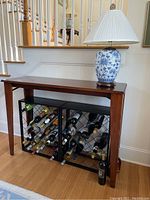 Wooden console table against wall, blue and white porcelain lamp on top, two black metal wine racks loaded with wine bottles underneath.