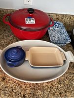 All five items displayed on countertop: red Dutch oven, orange oval dish, small blue pot, beige rectangular dish, speckled enamelware pan