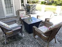 Four resin patio chairs arranged around a black square outdoor table on a pebble patio. Chairs have floral cushions.