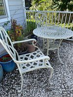 Three-piece white iron bistro set with two chairs and round table, placed on a patio with potted plants and railing in the background.