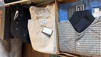 Image showing multiple shirts laid out in a box including two beige Alfani polos with tags, a black Polo shirt, light blue jeans, and a plaid Cambridge Classics shirt.