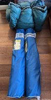 Green sleeping bag and two blue folding camp chairs in carrying bags arranged on wood floor.