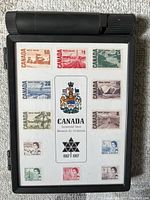 Front view of black collector box with full mint Centennial issue Canadian postage stamp set arranged in three columns and four rows on white backing.