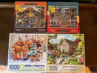 Four puzzle boxes on a wooden surface: two 1000-piece puzzles and two 500-piece puzzles from Bits and Pieces and Dowdle Folk Art