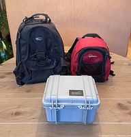 Photo of the two Lowepro backpacks, black and red, and the silver Pelican 1300 case in front of them on a wooden table.