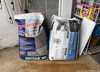 Photo shows two unopened bags: OptimaTrix polymeric sand and KRETE parging mix, placed on a floor near a metal rack.