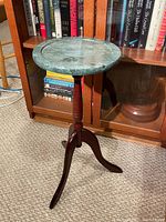 Green marble topped side table with a wooden three-legged base placed on carpet in front of a bookcase.