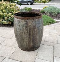 Metal barrel standing outdoors on stone patio, displaying full height and riveted panel construction.