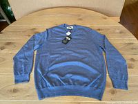 Full front view of blue men's sweater laid flat on wooden table, showing V-neck and ribbed cuffs and hem