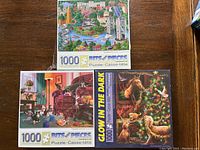 Three unopened Bits and Pieces 1000-piece puzzles stacked on a wooden surface showing the covers with detailed artwork.