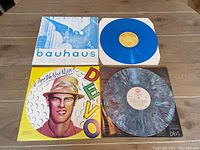 Two vinyl albums placed on a wooden surface: Bauhaus Bela Lugosi's Dead in blue vinyl with matching cover and Devo Are We Not Men in multicoloured vinyl with colorful cover.