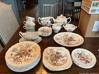 Full set of plates, cups, and serving items arranged on wood table showing floral pattern and scalloped edges.