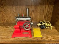Front and right side view of the Weeden No. 670 horizontal electric steam engine on a wooden shelf, showing red painted base, silver boiler, flywheel and chimney, with attached cord and control switch lying beside it.