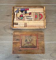 Top view of the wooden blocks neatly arranged inside the original wooden box with sliding lid. The box lid is placed beside it, showing vintage artwork of children playing with blocks.