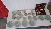 Overview of celadon porcelain dish set and Oneida Queen Bess silverplate flatware in wooden case on white table.