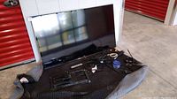 Samsung smart TV leaning against a white partition, mounted on a black protective moving blanket. Various accessories and cables laid out beside it.