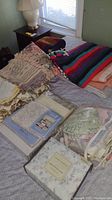 Photo showing assorted layered blankets and packaged sheet sets on bed including striped fleece, tribal style woven blanket, and floral sheet set packages