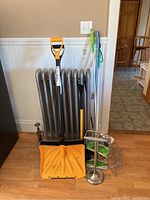 Two snow shovels, two Swiffer sweepers, broom, metal soap holder, and toilet paper holder arranged against a radiator and wall.
