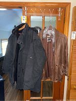Two coats hanging on a wooden door, a dark cloth Hardy Amies London coat with leather trim and a brown Aananda leather coat