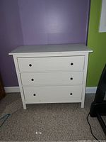 Front view of a white wooden dresser with three drawers against a purple and green wall.