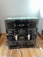 Front view of black wooden Asian-style end table with glass top, one drawer, two doors, and carved painted figures and landscapes.