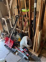 Photo showing clustered yard tools including shovels, rakes, pick, weed trimmer, and bird netting in basement corner.