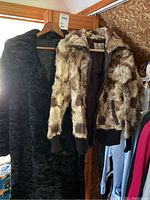 Two fur coats hanging on wooden hangers on a stand displaying entire coats, one black and textured and one light brown patterned.